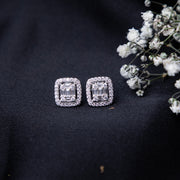 A pair of sterling silver cushion-shaped earrings with intricate detailing, displayed on a dark fabric surface alongside some small white flowers.